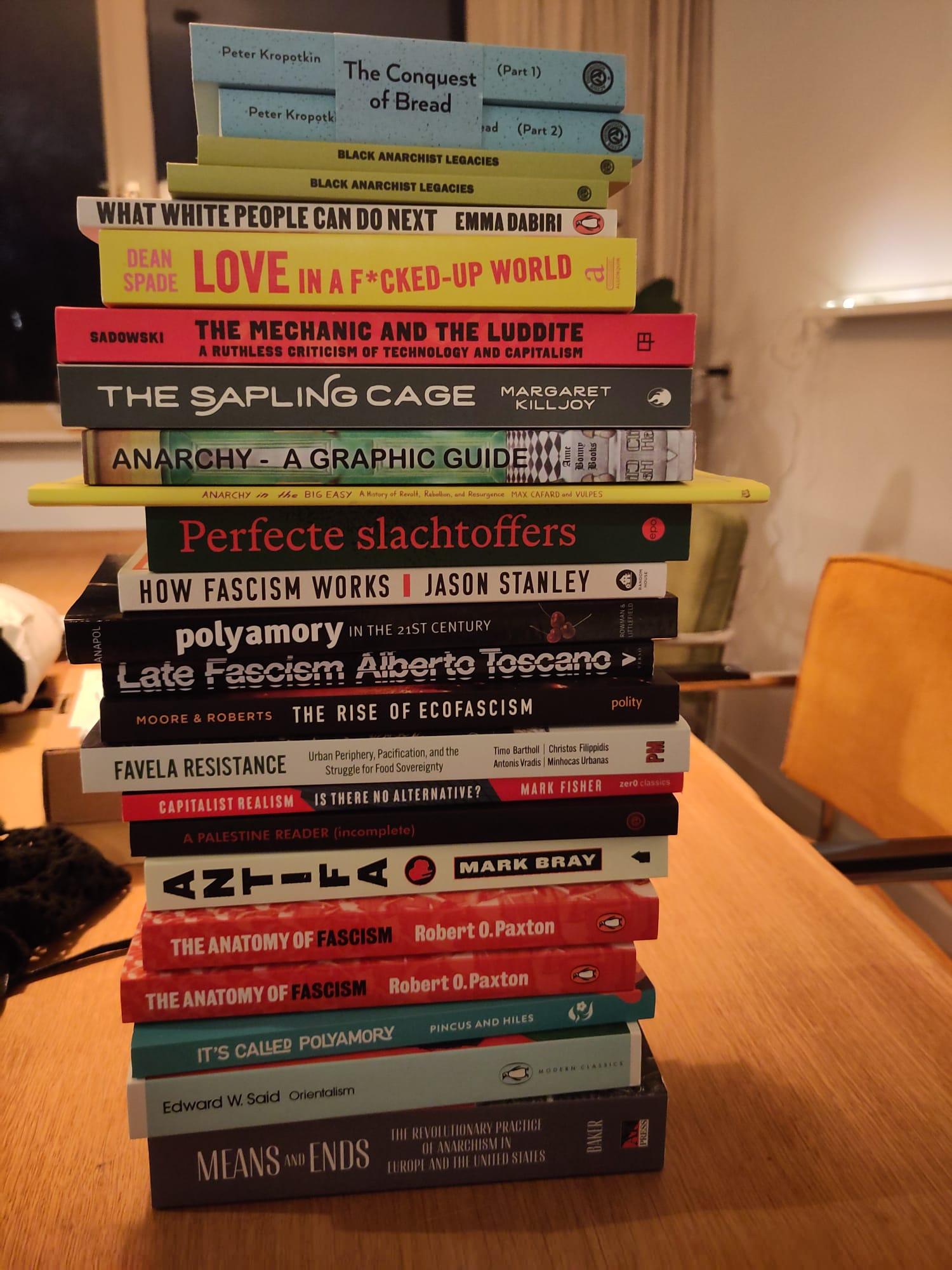 A pile of 24 books over a wooden table, with an orange chair and a window in the background.